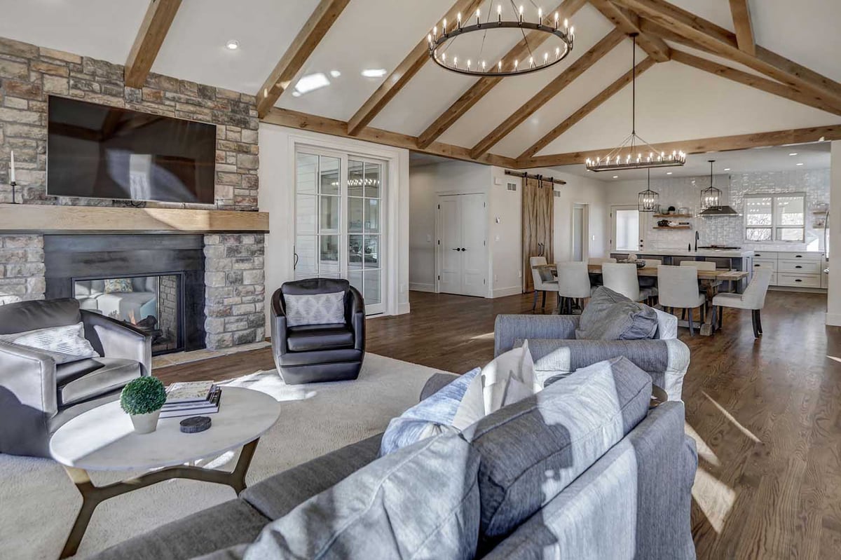 A bright and airy open-concept living room with exposed wooden beams, a stone fireplace, and a view into the adjoining dining area and kitchen.