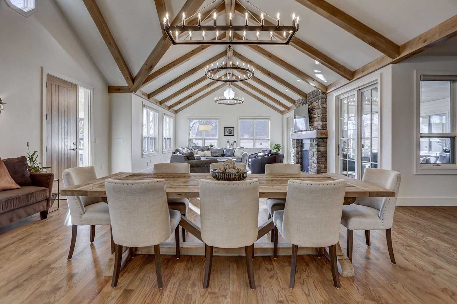 A spacious open-concept living and dining area featuring exposed wooden beams, a large rustic dining table, and a cozy stone fireplace.
