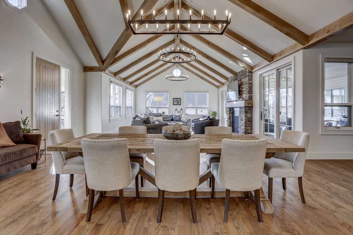 A spacious open-concept living and dining area featuring exposed wooden beams, a large rustic dining table, and a cozy stone fireplace.