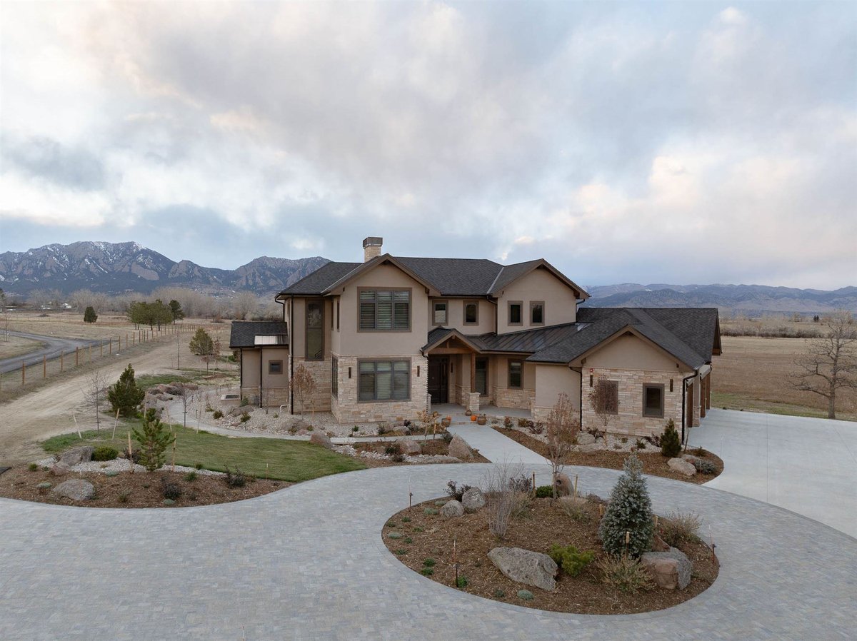 exterior view of a large home remodel with mountain views, warm tan/brown stone exterior, and a large gray stone driveway
